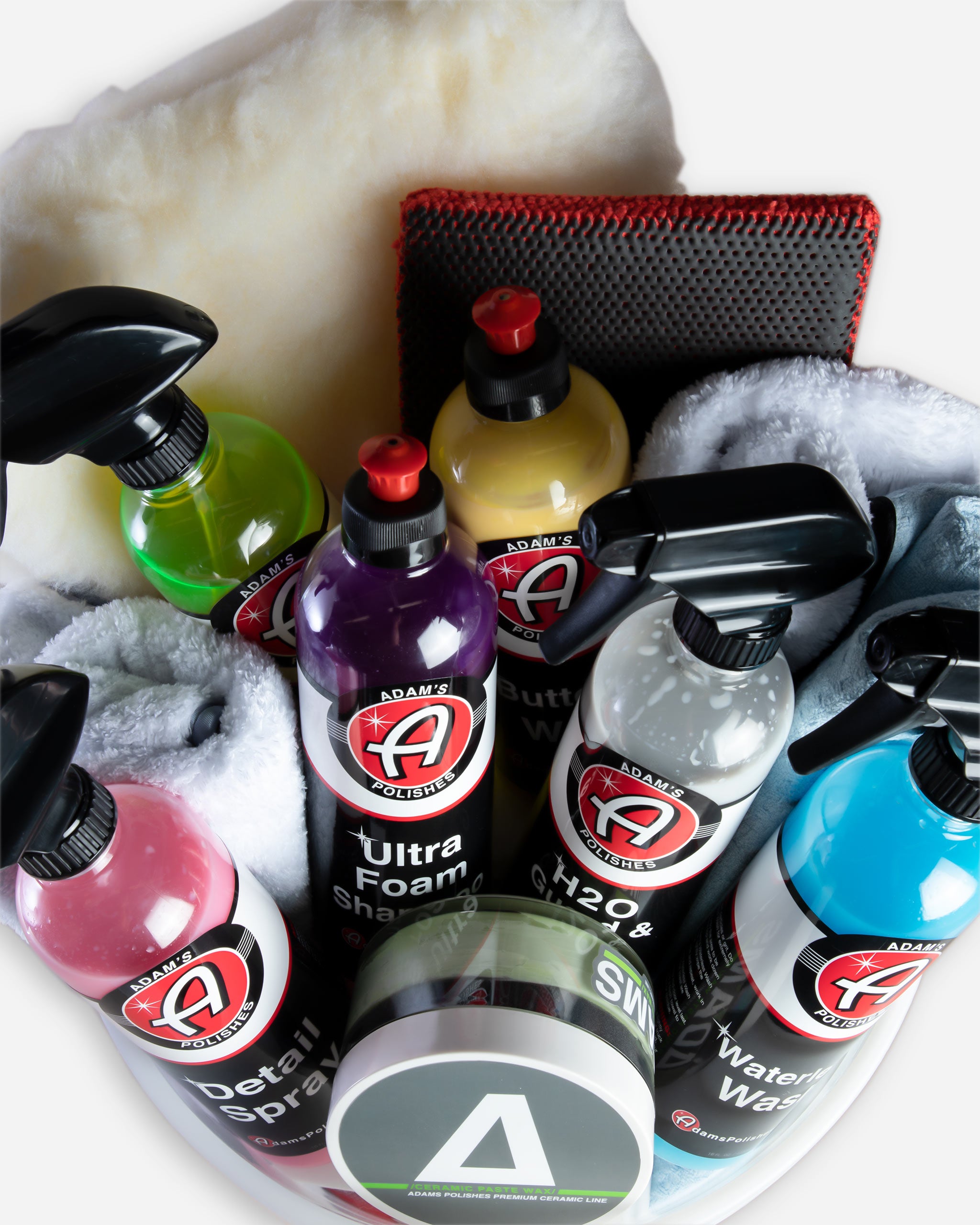 Alt text: Assortment of Adam's car care products displayed prominently, including bottles of brightly colored Ultra Foam Shampoo, Detail Spray, and H2O Guard & Gloss, alongside microfiber towels and washing pads, organized neatly inside a mystery bucket