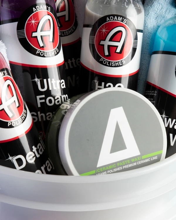 Alt text: Close-up view of Adam's Mystery Bucket filled with various car care products including bottles labeled "Ultra Foam Shampoo" and "H2O Guard & Gloss," and a container of "Ceramic Paste Wax," prominently displaying the Adam