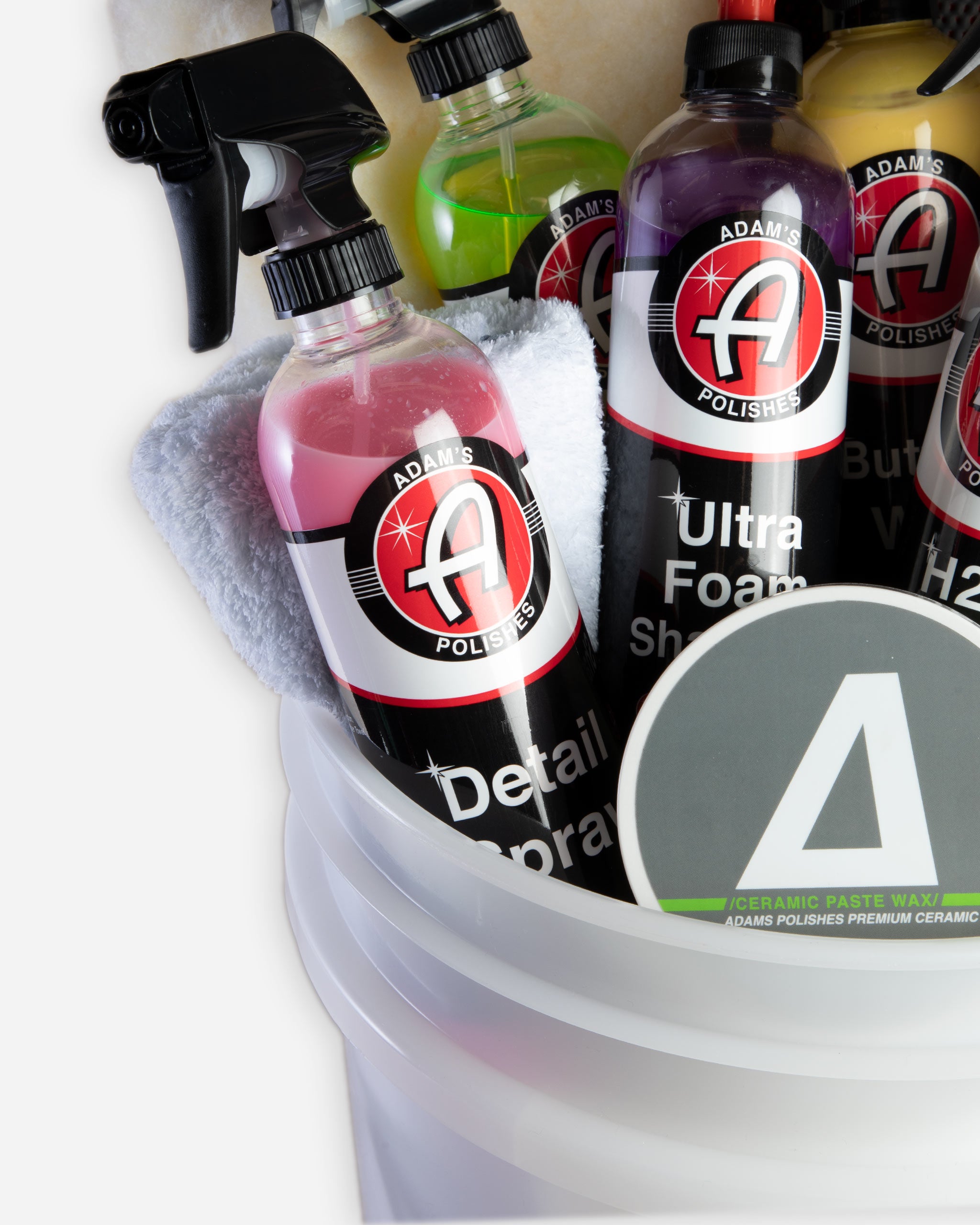 Alt text: Variety of car care products including bottles of Adam's Polishes, Ultra Foam Shampoo, H2O Guard & Gloss, and Detail Spray, prominently displayed in a transparent white bucket with an Adams Polishes logo, arranged with a micro
