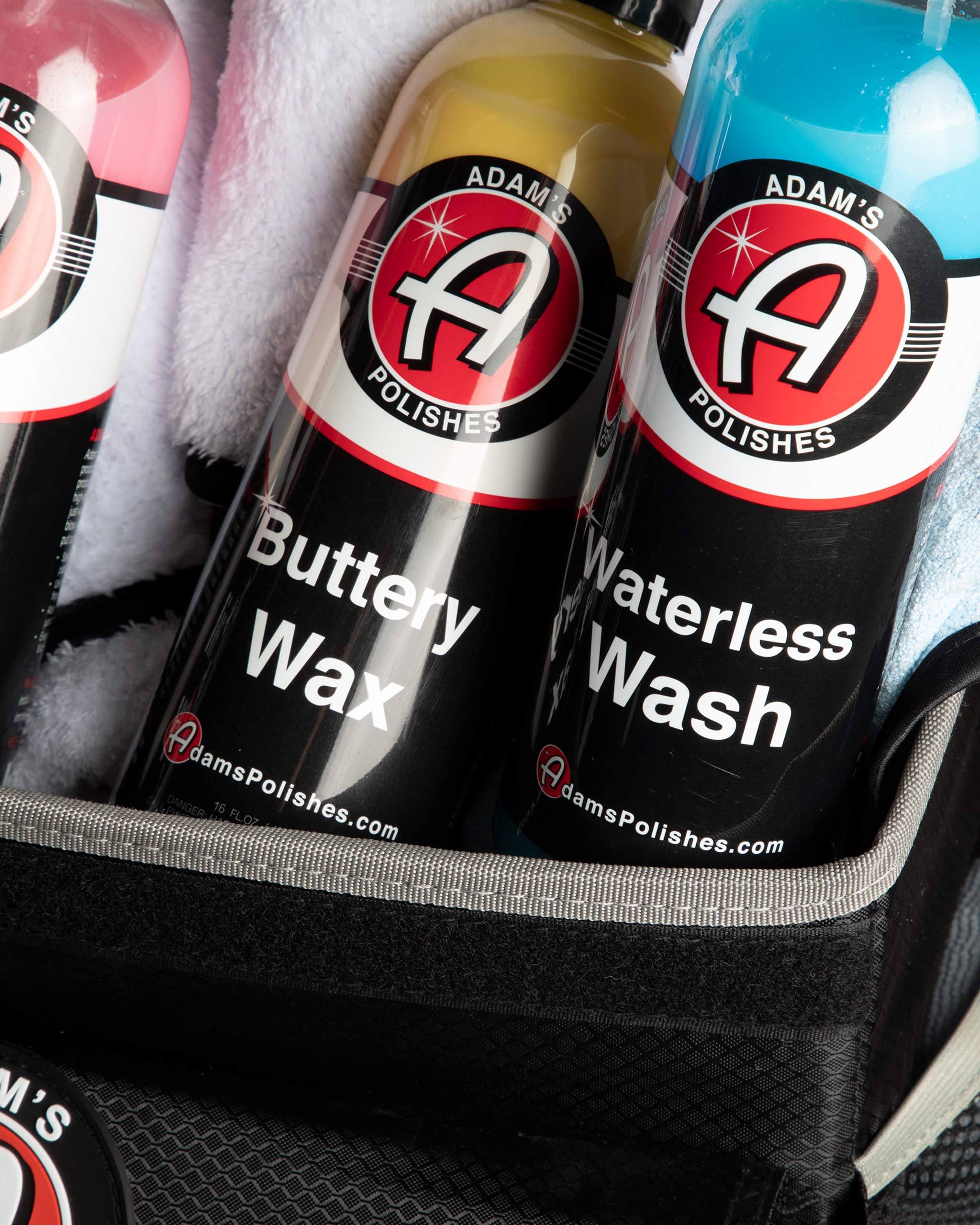 Alt text: Close-up view of Adam’s car care products in a trunk organizer, featuring bottles of Buttery Wax and Waterless Wash with prominent red and black branding, nestled in a black fabric organizer with visible stitching and a plush white towel.